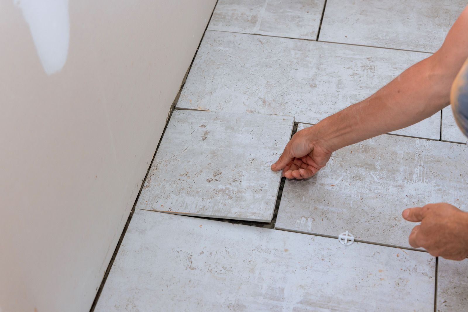 Hands adjusting a lifted white floor tile next to a wall during installation.
