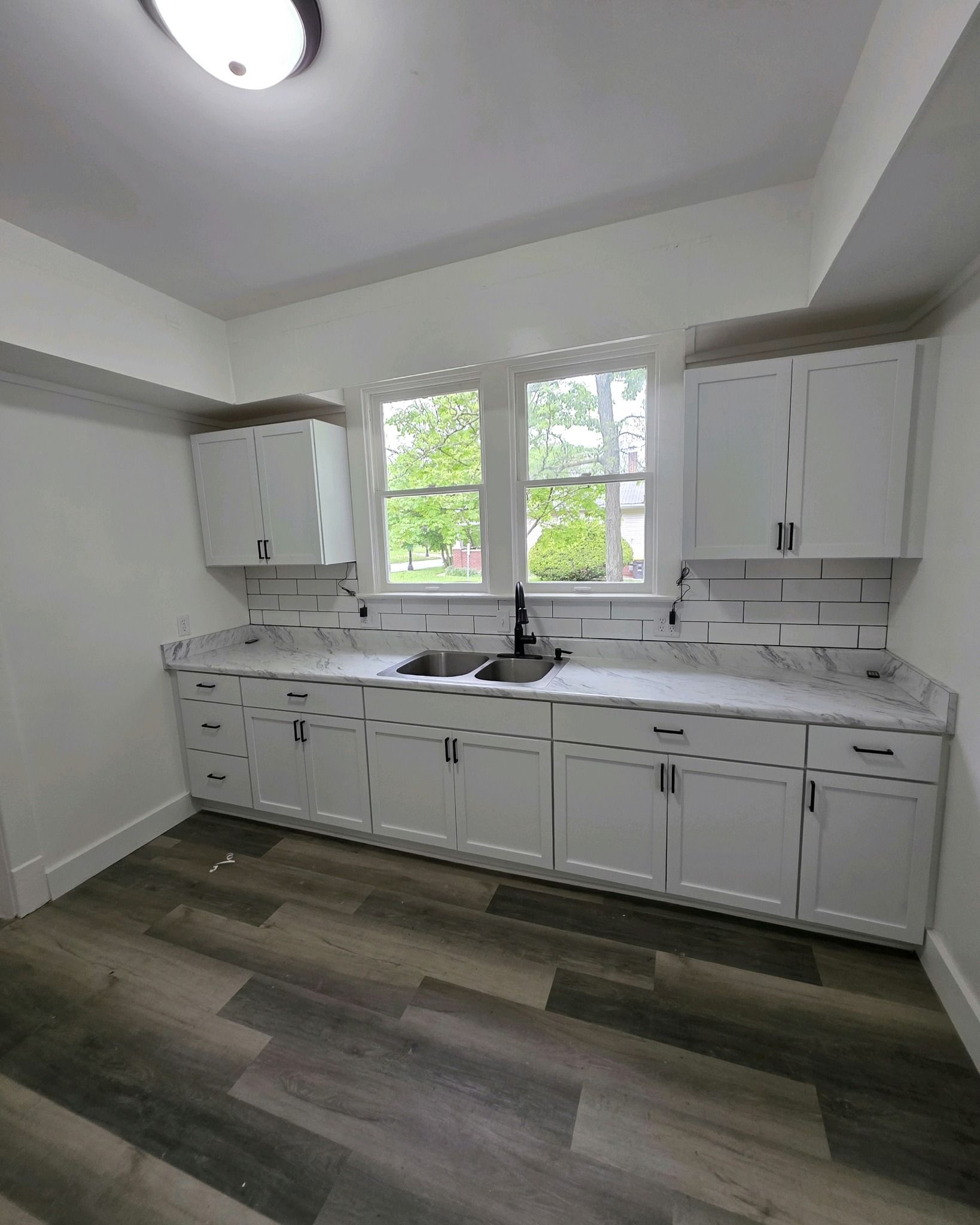 Bright white kitchen with cabinets, central window, sink, and gray wood-style floor.