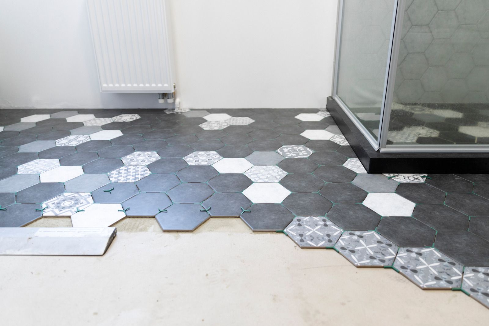 Hexagonal floor tiles scattered across a room, with gray, white, and blue pieces near a radiator and glass shower.
