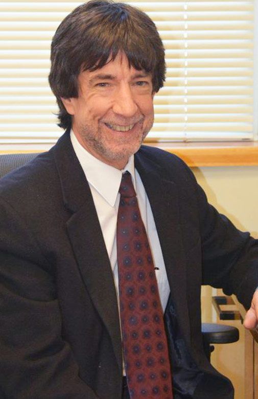Smiling man in a suit and tie seated indoors beside a window with blinds.