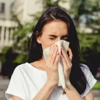 Woman in a white shirt outdoors, eyes closed, blowing her nose into a tissue.