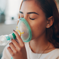 Woman using a nebulizer mask with eyes closed in a bright indoor setting
