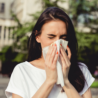 Woman in a white shirt outdoors, eyes closed, blowing her nose into a tissue.