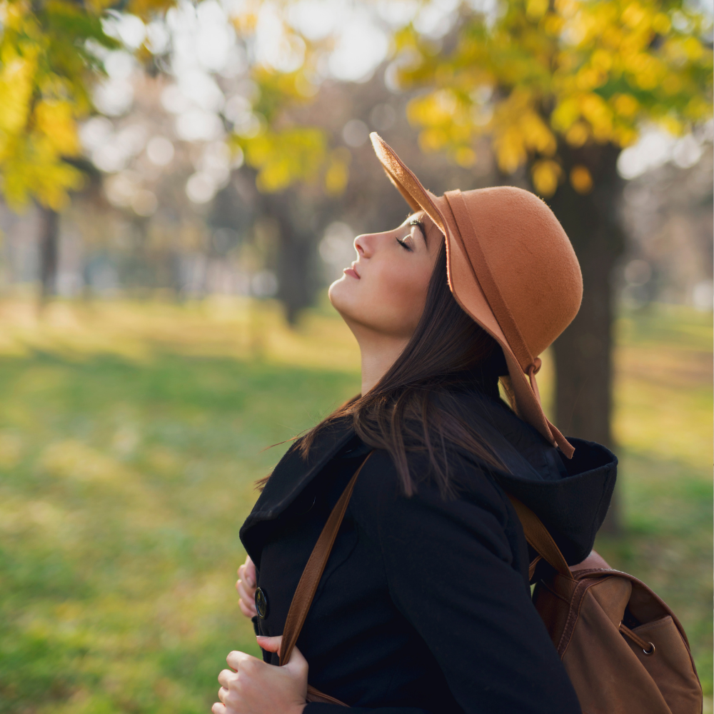 Woman in a brown hat outdoors in a park, looking up among autumn trees, carrying a backpack