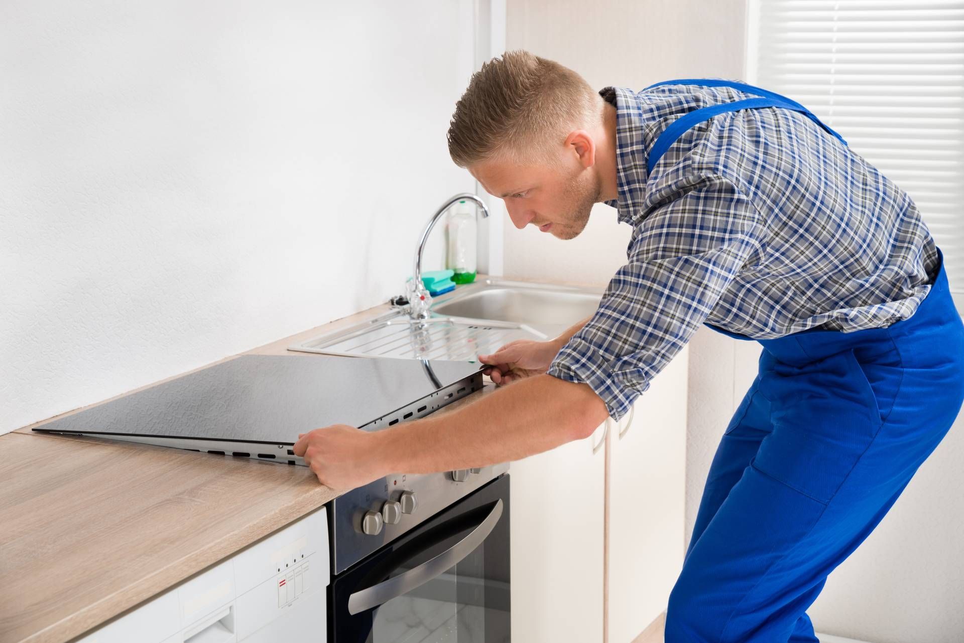 A man in blue overalls installing a black stovetop in a kitchen.