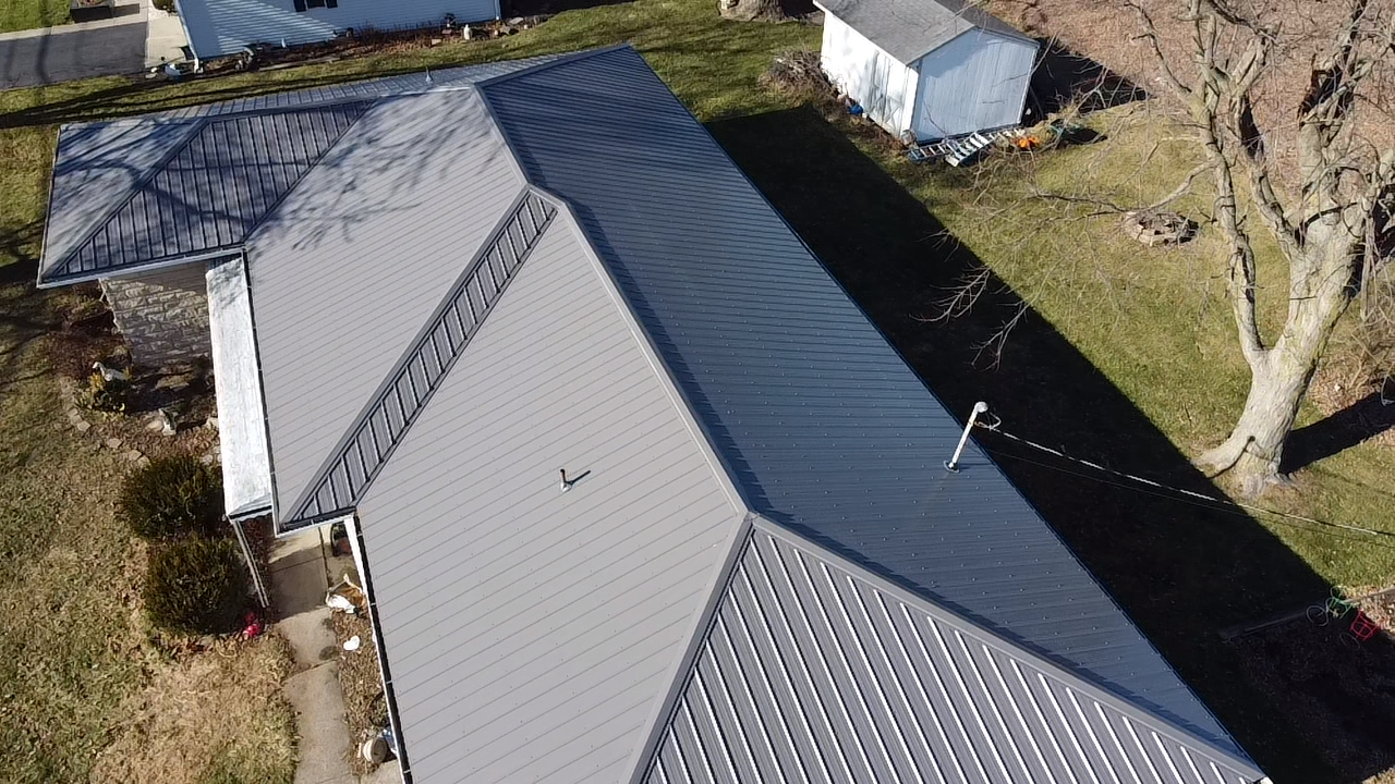 Aerial view of a house with a gray metal roof, surrounded by grass and a tree.