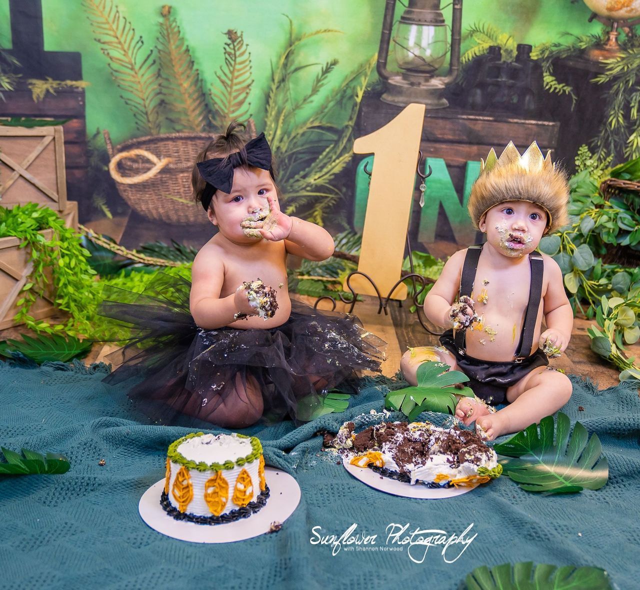 Two babies are sitting on a blanket eating cakes.