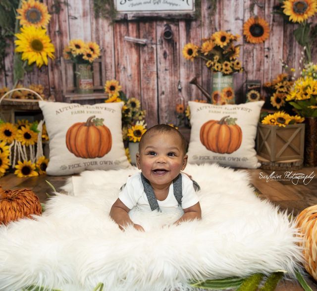 A baby is laying on a white blanket with pumpkins and sunflowers in the background.