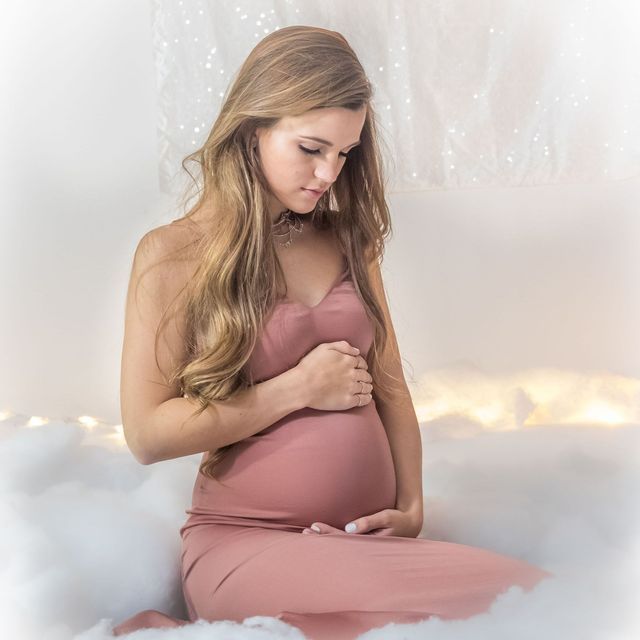 A pregnant woman in a pink dress is sitting on a bed holding her belly.