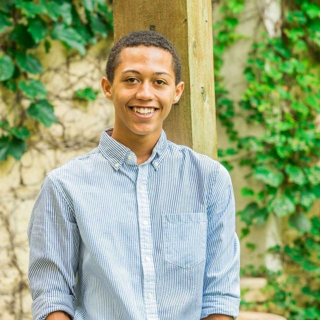 A young man in a blue striped shirt is smiling while leaning against a wooden post.
