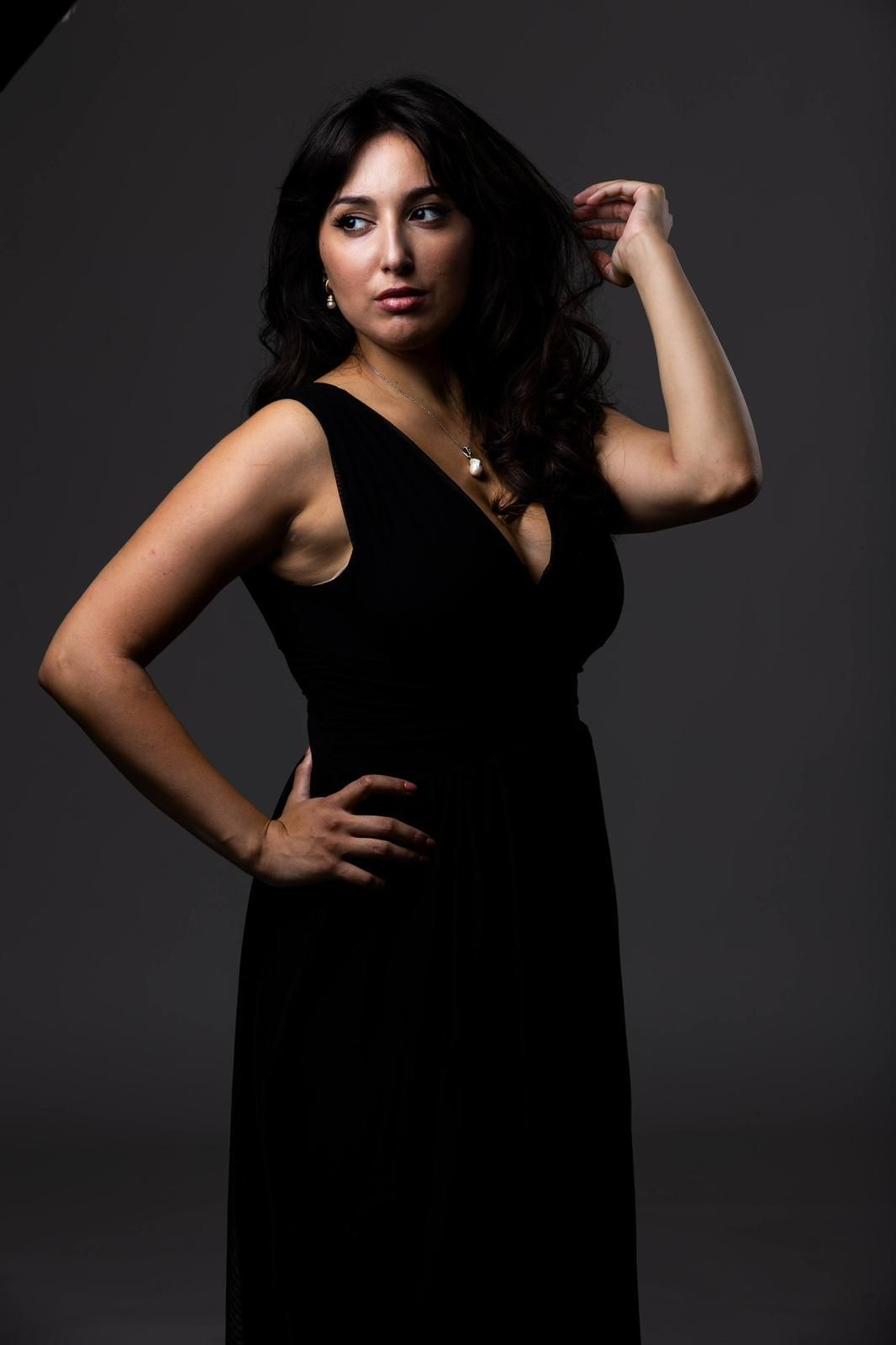 Donna in abito nero, con le mani nei capelli, in posa su uno sfondo grigio.