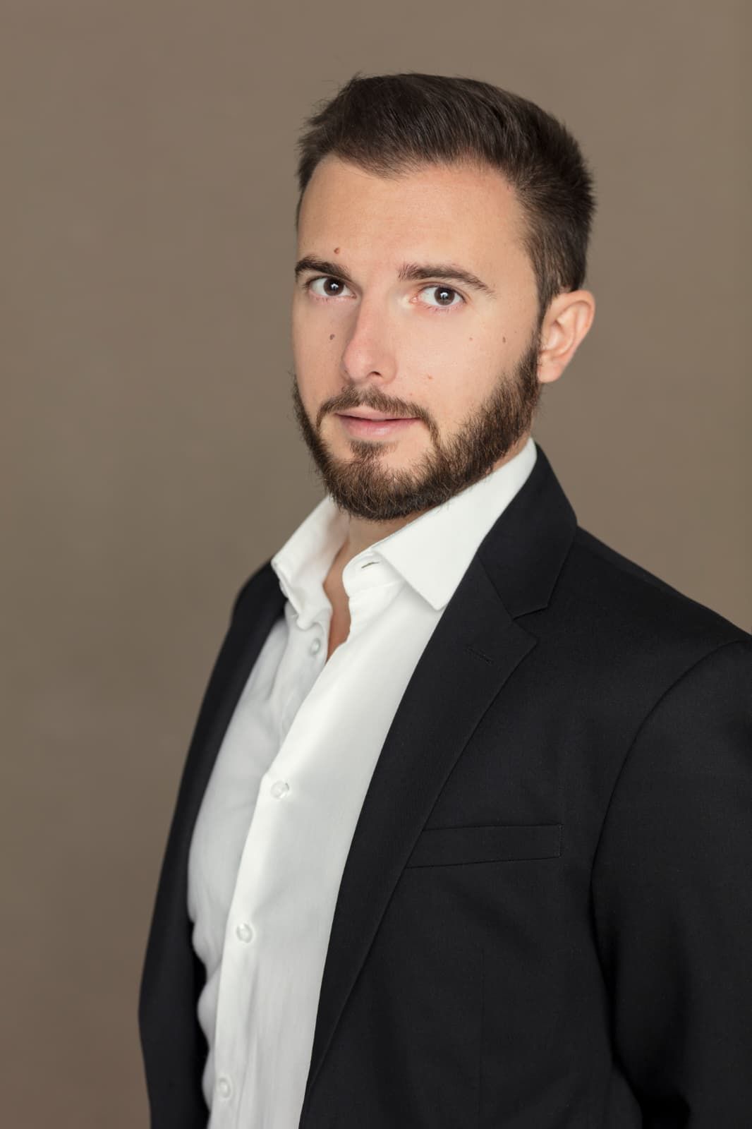 Uomo con blazer nero e camicia bianca, che guarda la telecamera su uno sfondo neutro.
