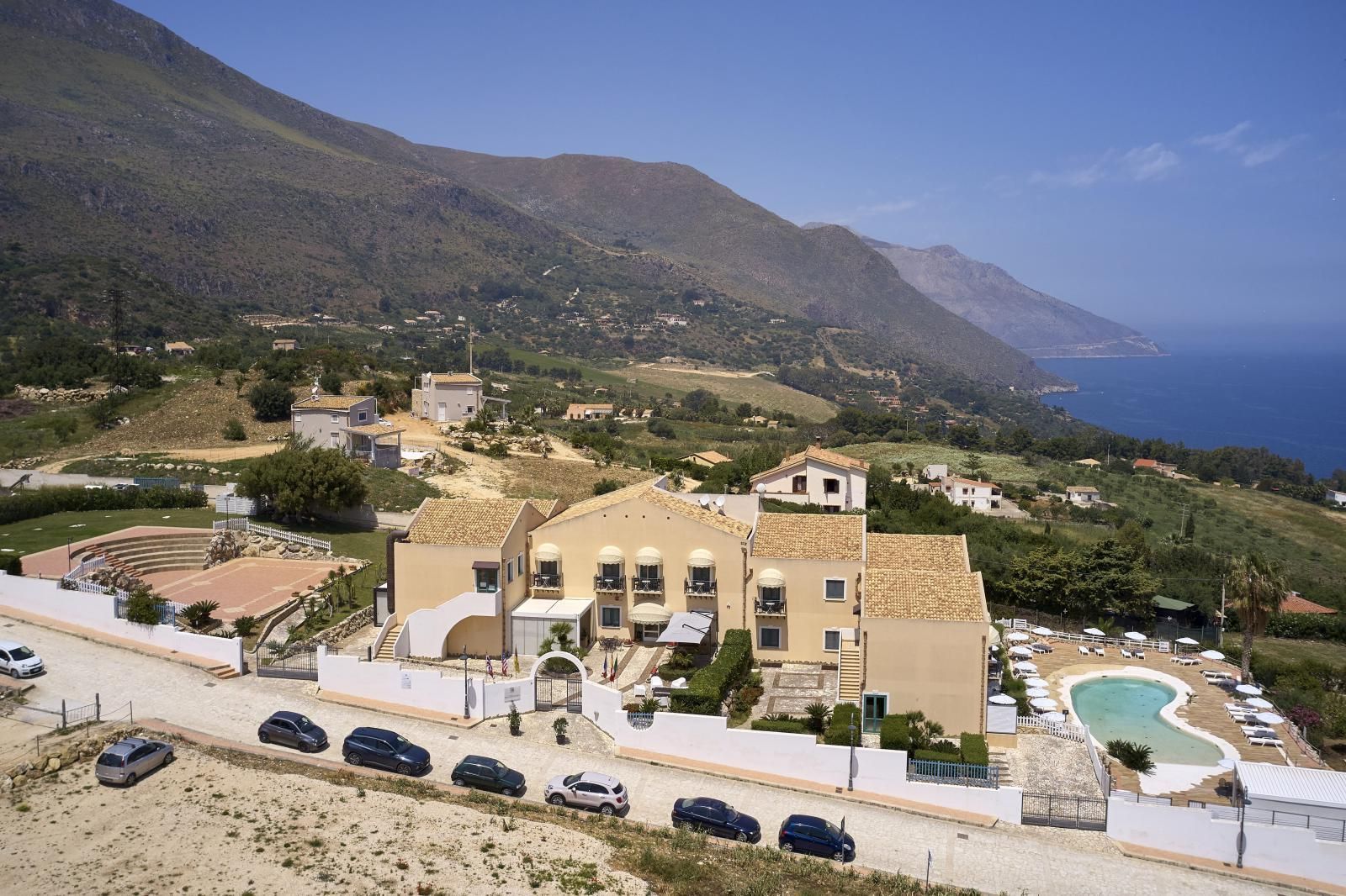Complesso alberghiero con piscina, vista sull'oceano e sulle montagne. Auto parcheggiate sulla strada.