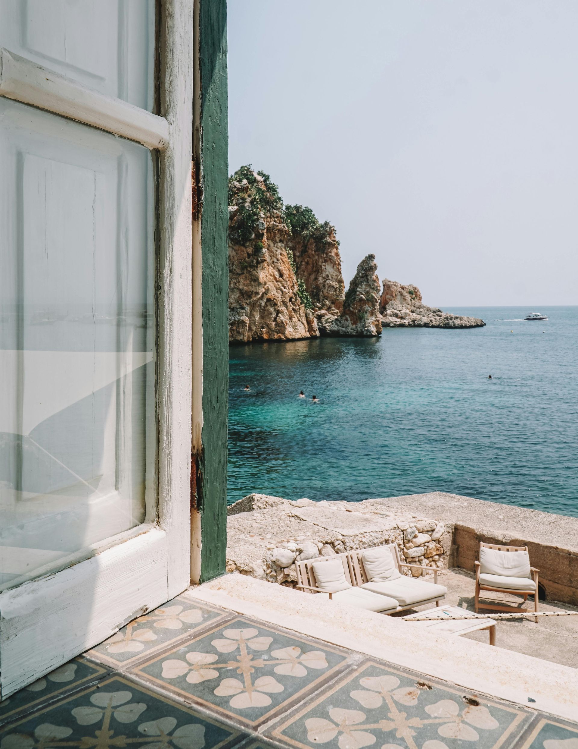 Vista dalla finestra di una costa rocciosa, acqua turchese, lettini e pavimento piastrellato blu.
