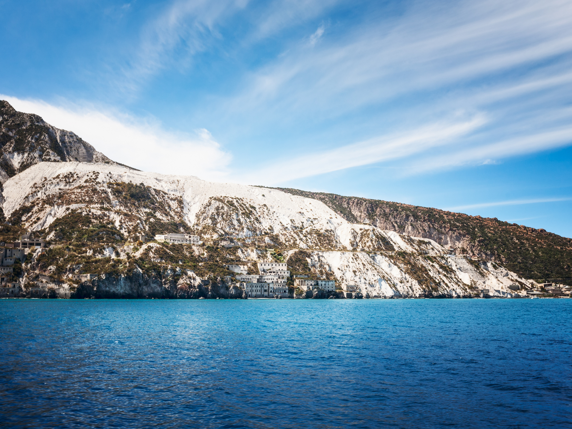 Cava di marmo bianco sul fianco di una montagna, sopra l'acqua azzurra, sotto un cielo luminoso e nuvoloso.
