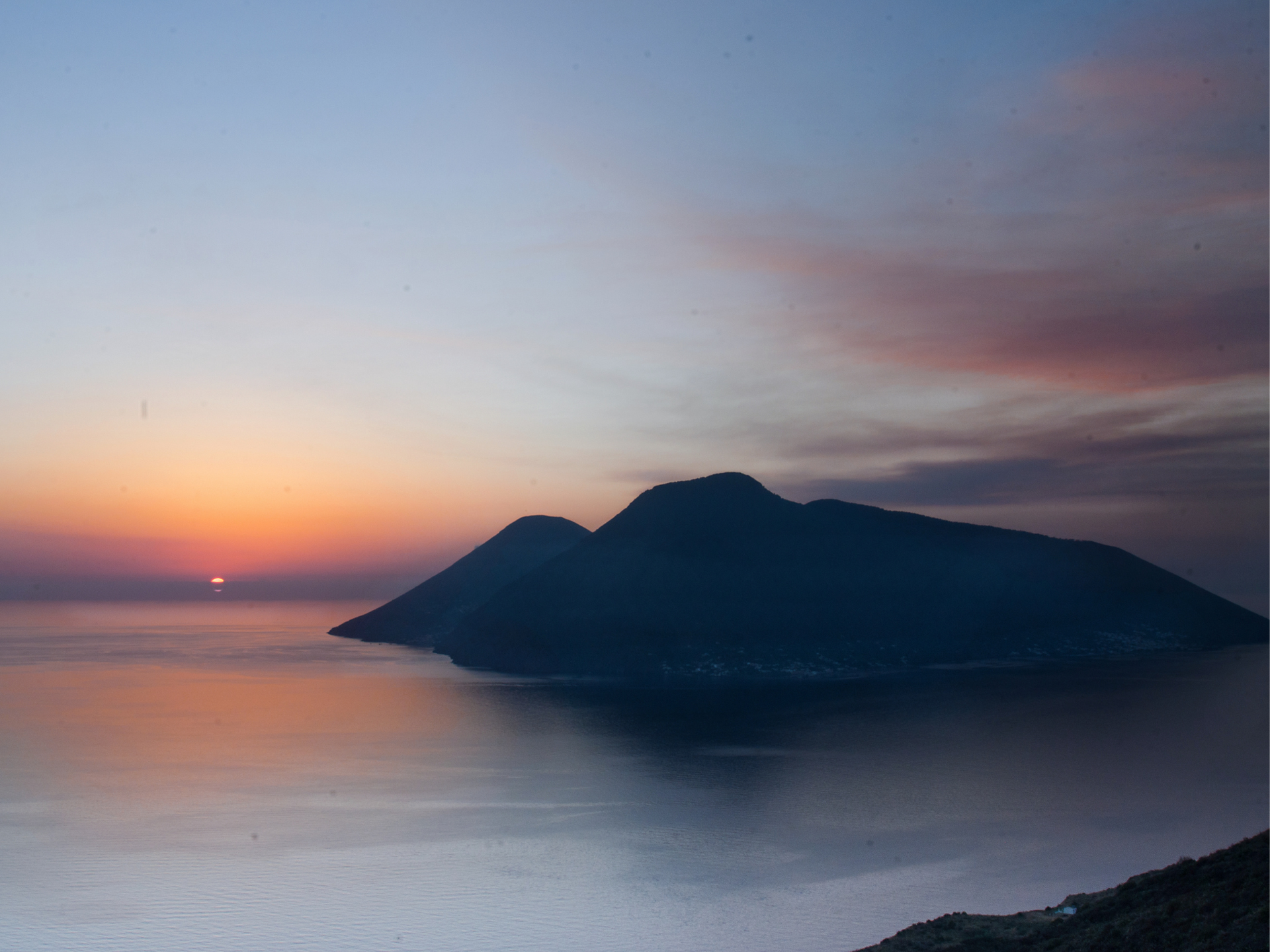 Tramonto su acque calme, isola in silhouette. Le sfumature arancioni e viola del cielo si riflettono nell'acqua.