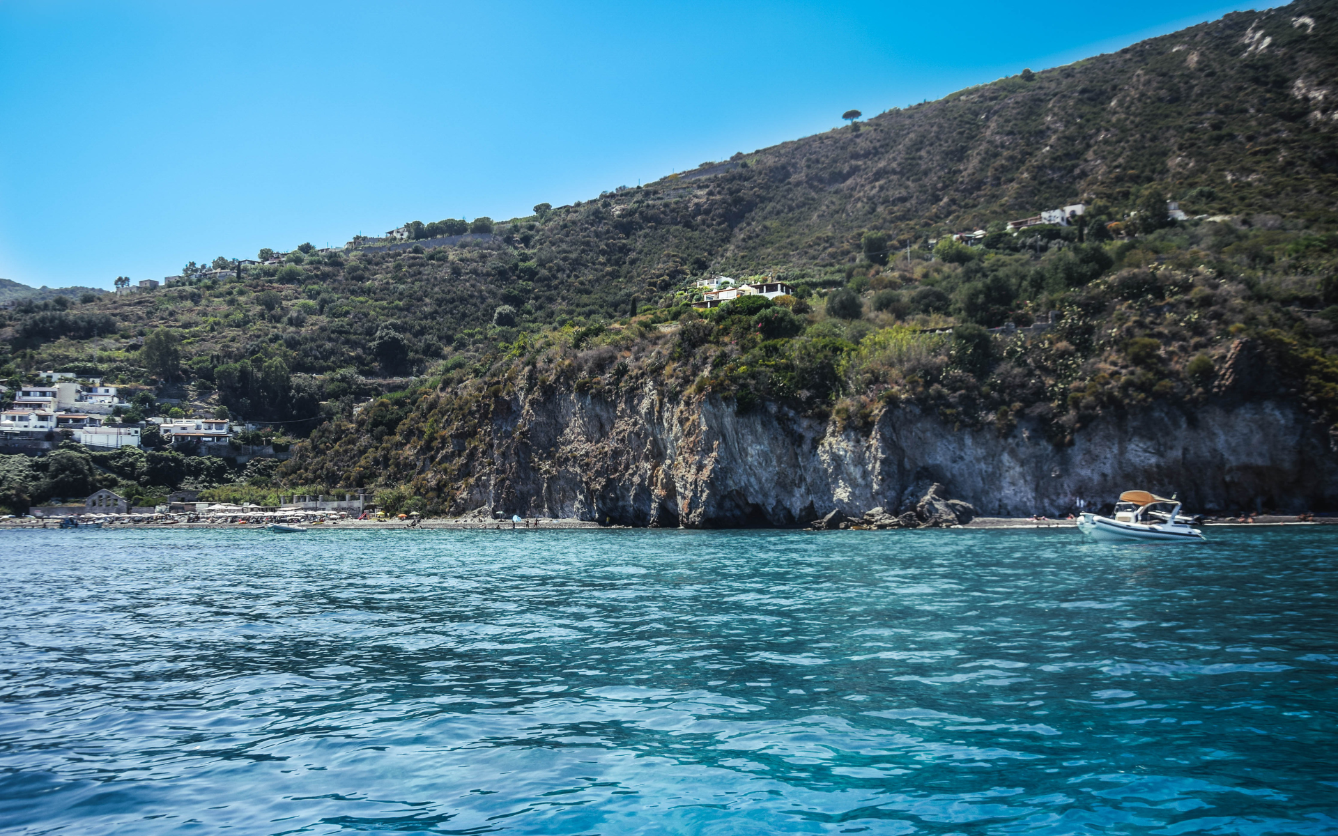 Isola con una città sulla costa, parzialmente coperta dalle nuvole. Cielo azzurro, acqua e fiori rossi in primo piano.