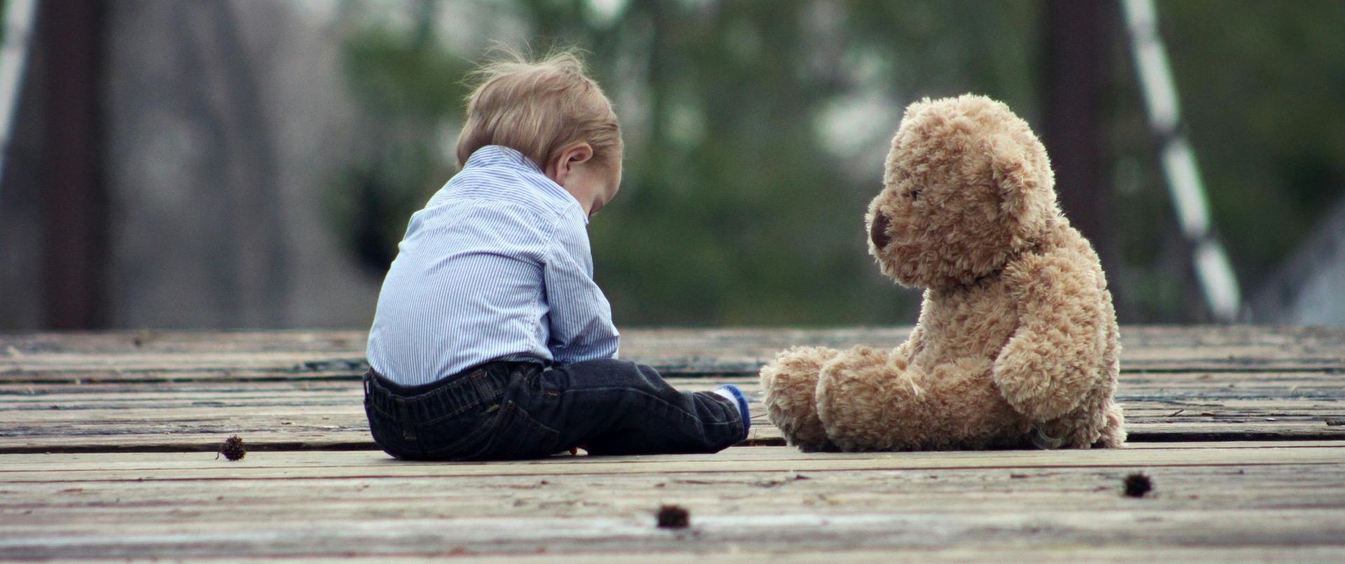 A little boy is sitting next to a teddy bear on a wooden deck.