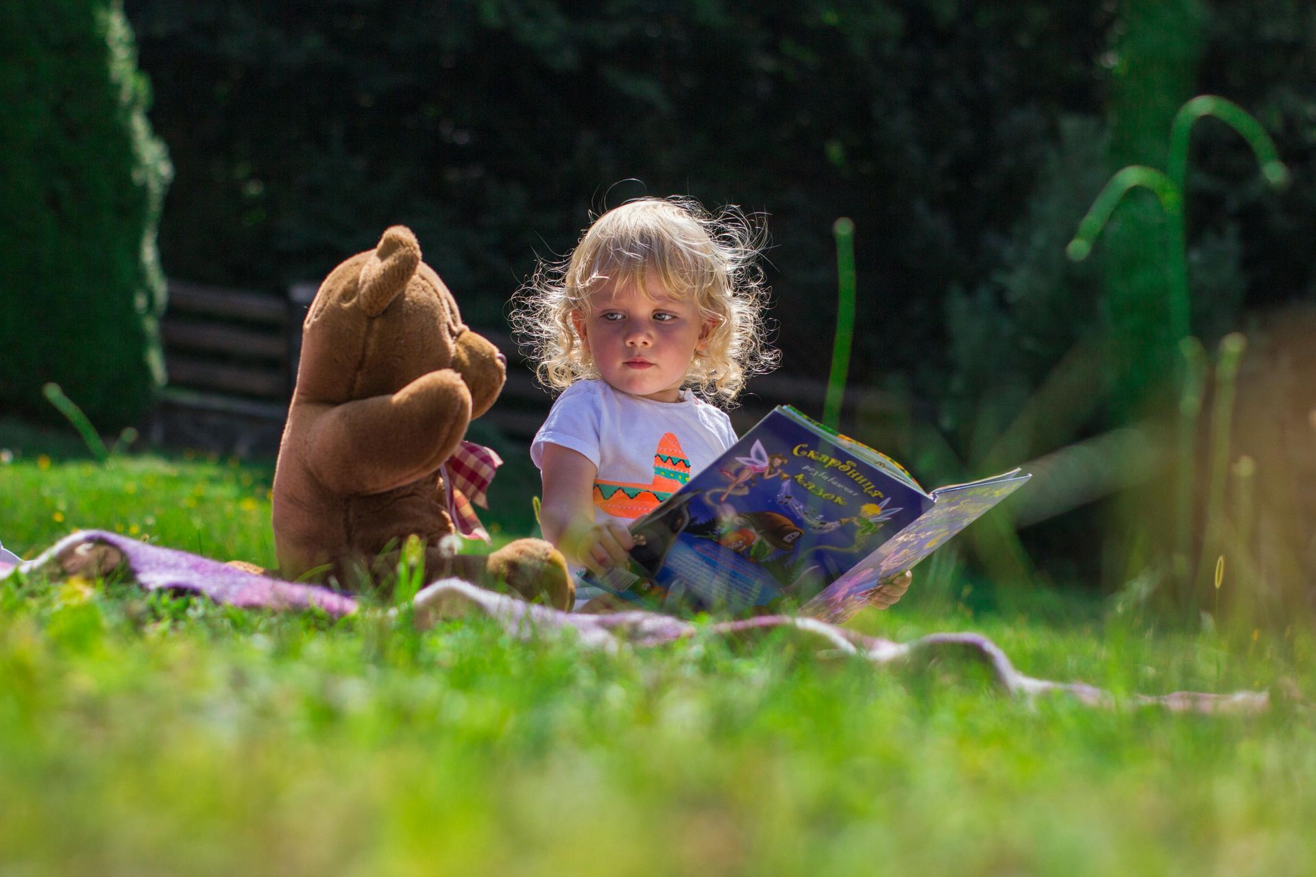 A little girl is sitting in the grass reading a book to a teddy bear.