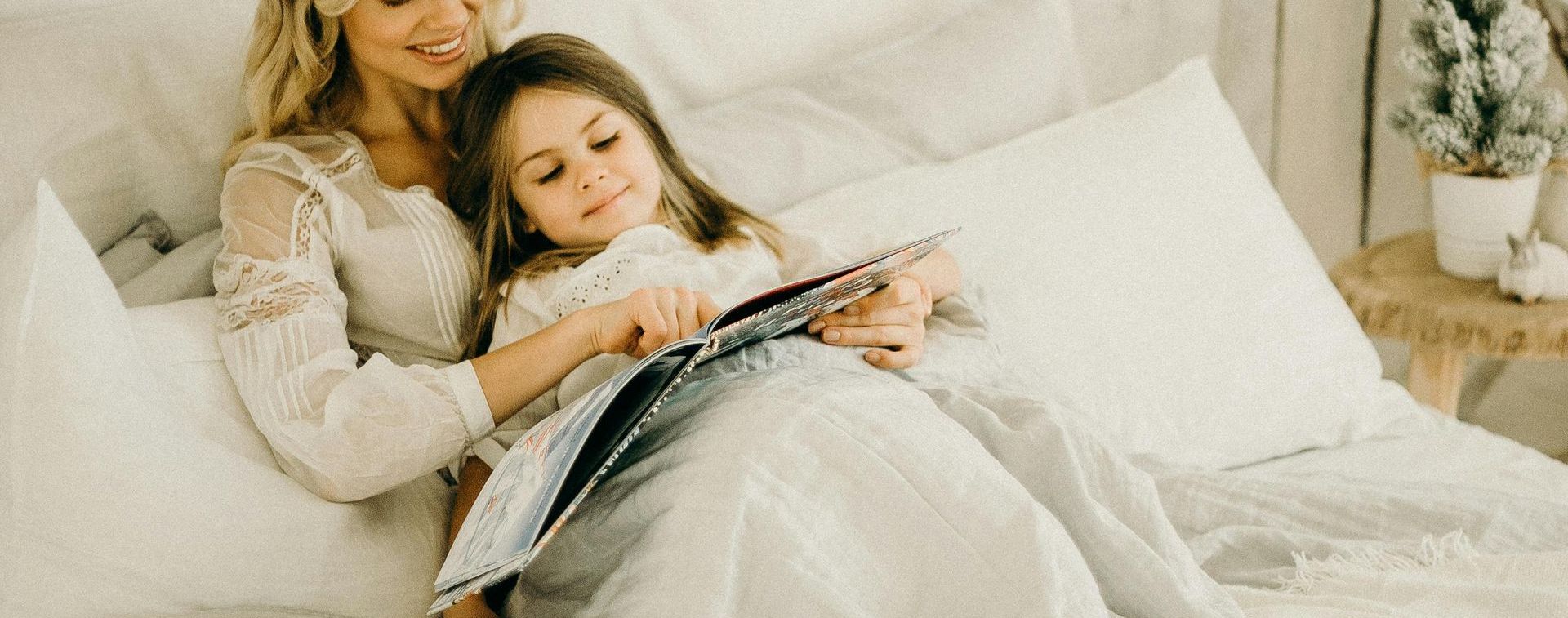 A woman and a little girl are sitting on a bed reading a book.