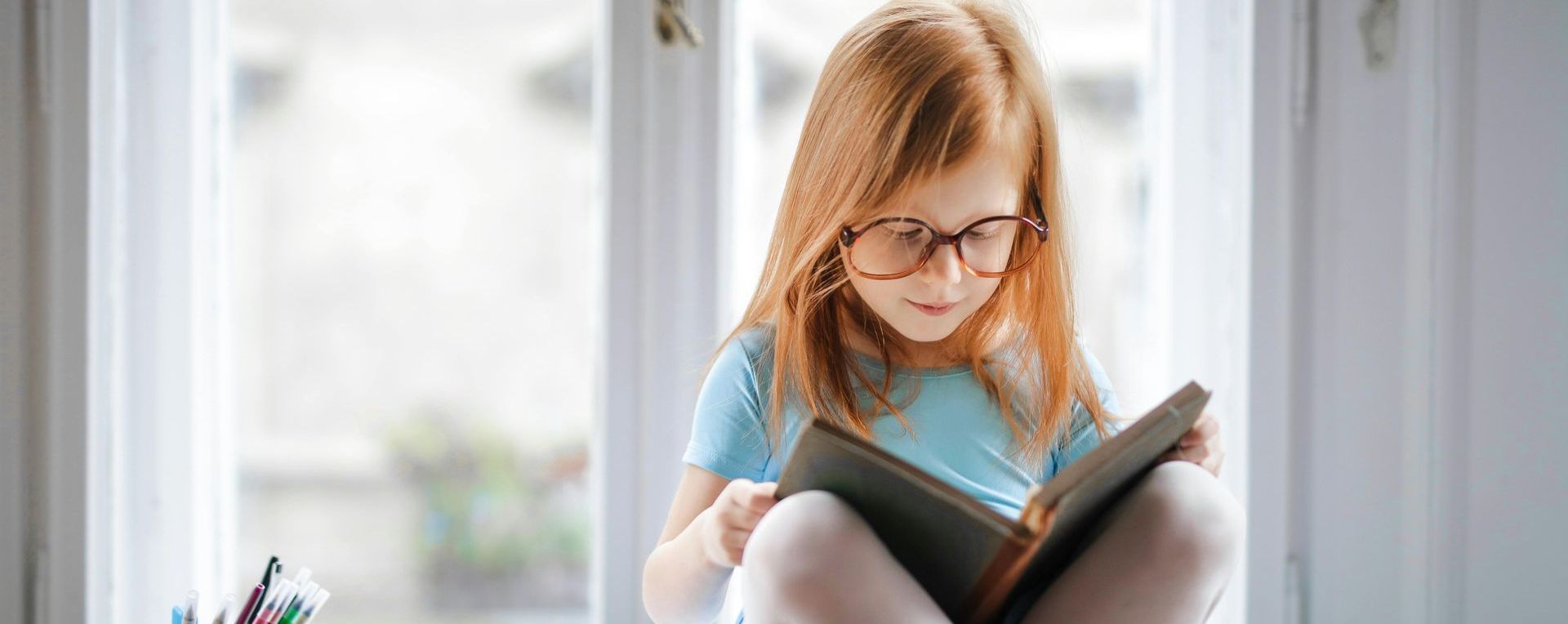 A little girl is sitting on a window sill reading a book.