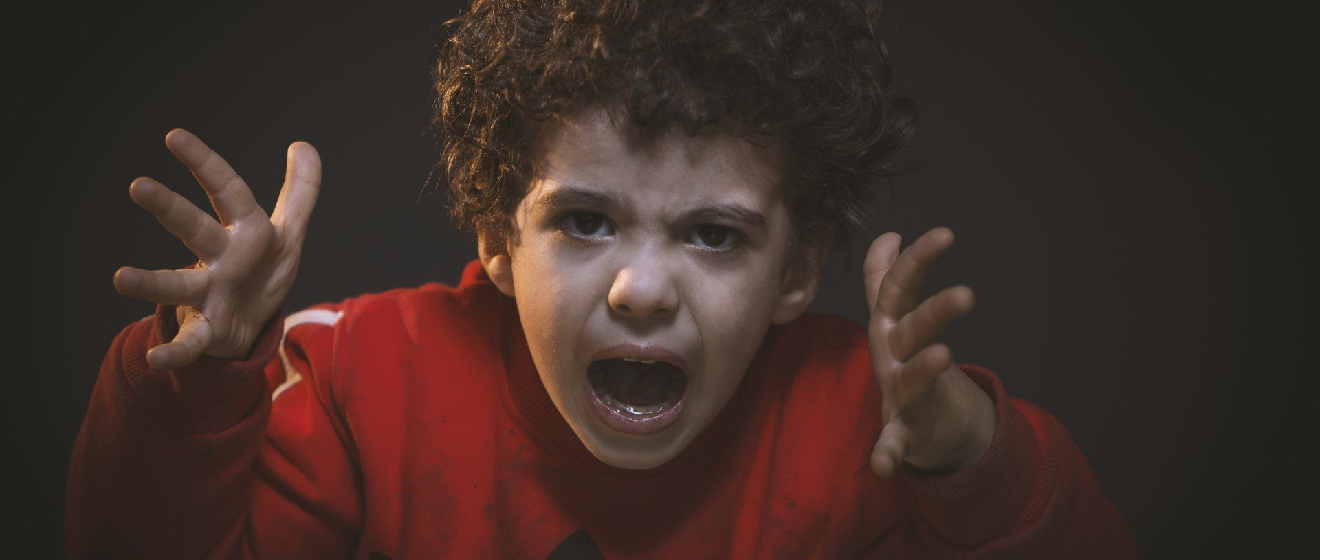 A young boy with curly hair is making a scary face with his hands in the air.