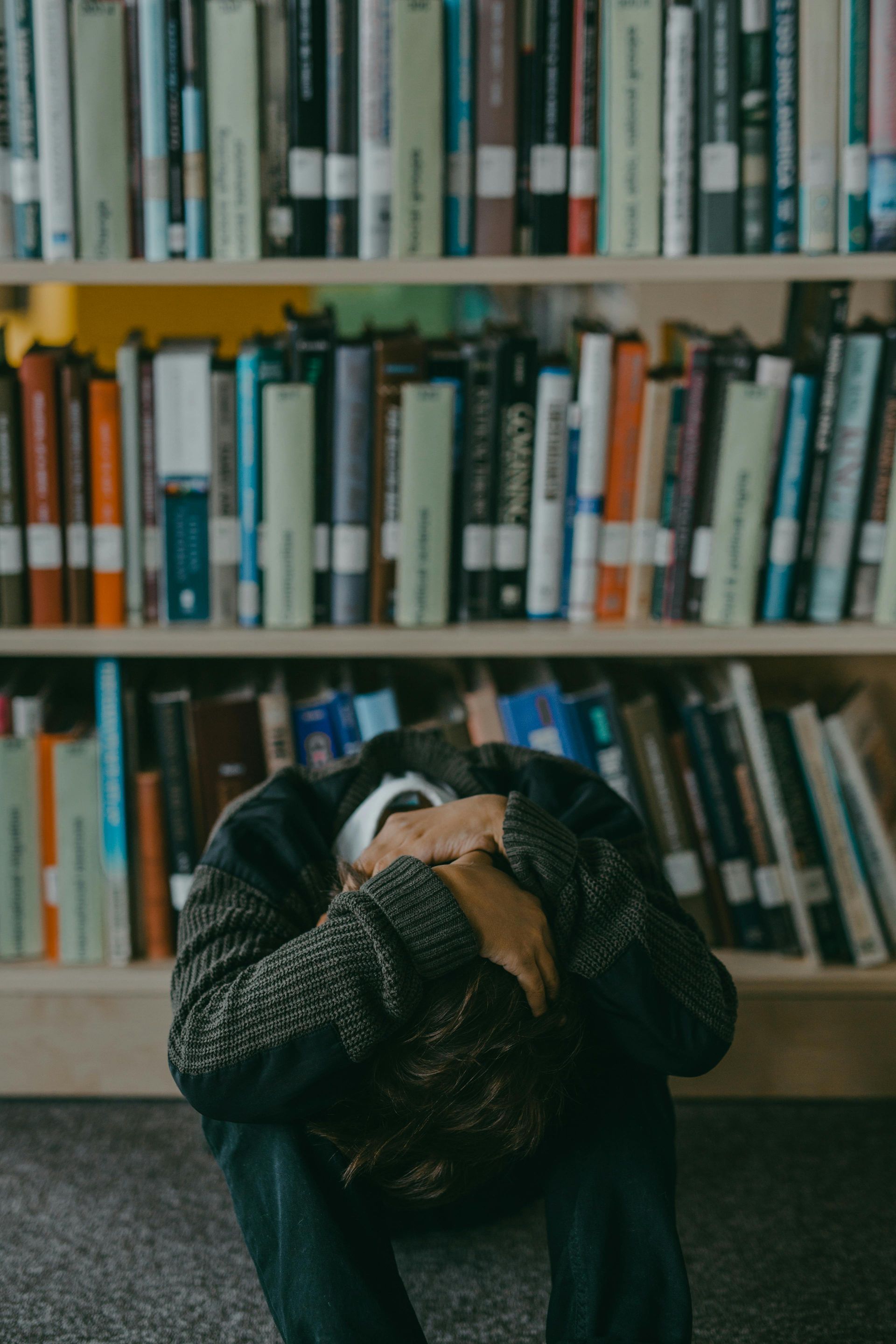 A man is sitting on the floor with his head in his hands in front of a bookshelf.