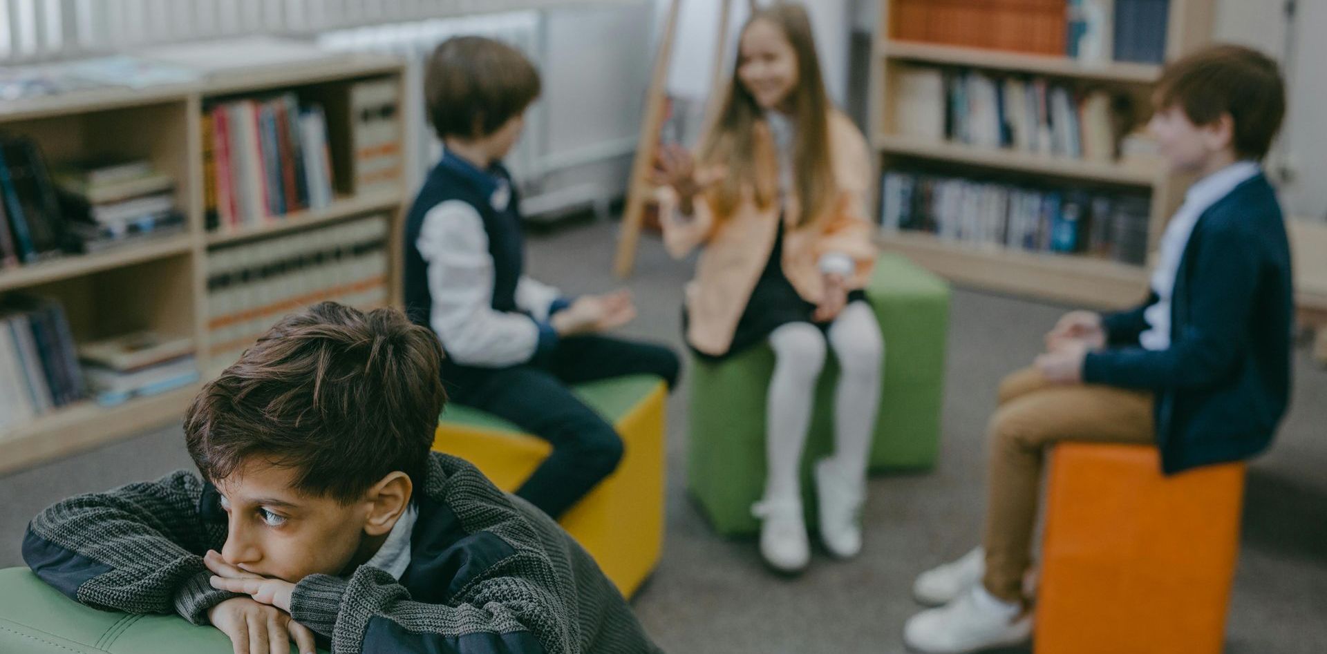 A group of children are sitting on ottomans in a library.
