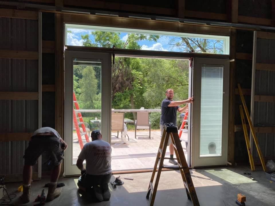 Three people installing glass doors in a building. The doors open to a deck and trees.