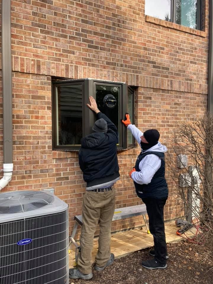 Two people installing a window on a brick building exterior; one uses orange gloves.