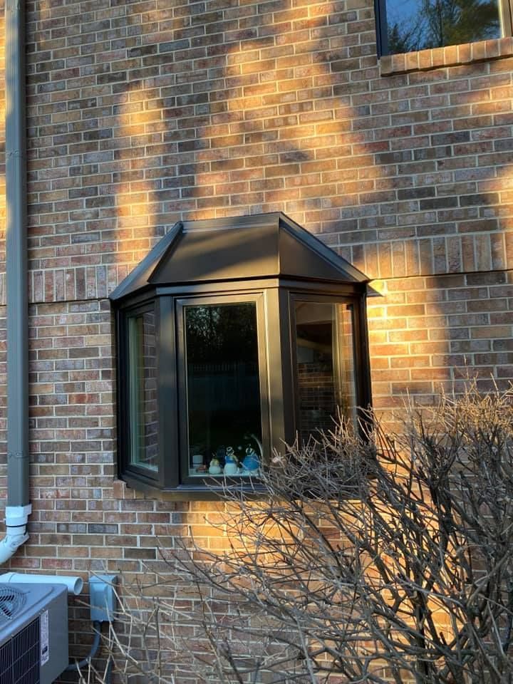 Brick building with a dark-framed bay window. Window has a dark roof. A bush is in the foreground.