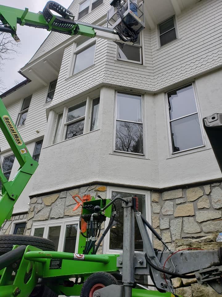 A lift platform with a person working on the windows of a white house with stone foundation.