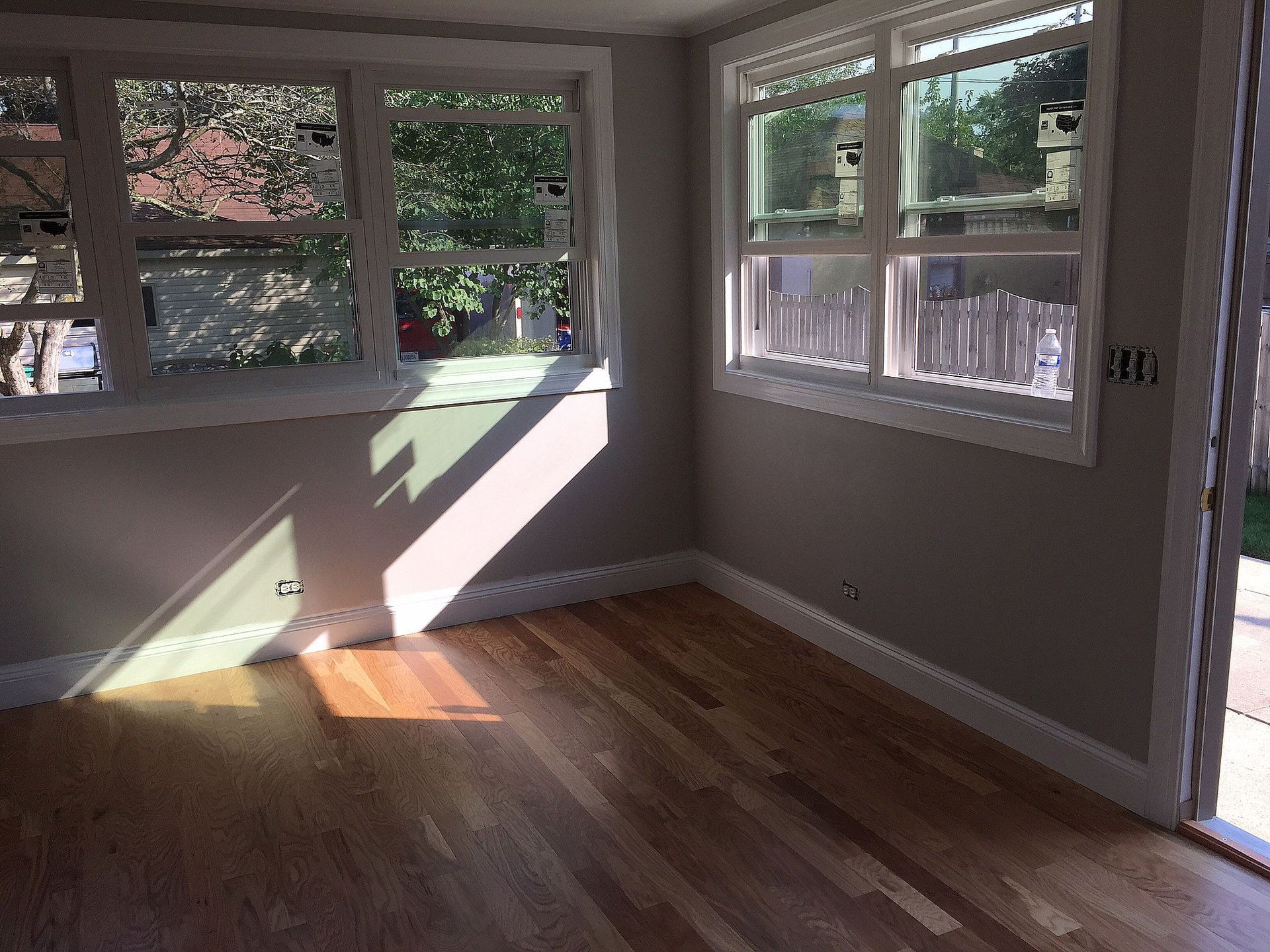 Sunlit room with wood floors, neutral walls, and multiple windows looking outside.