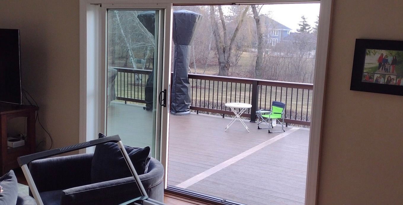 Sliding glass door open to a wooden deck with railing, trees, and small table. Inside shows a couch and a TV.
