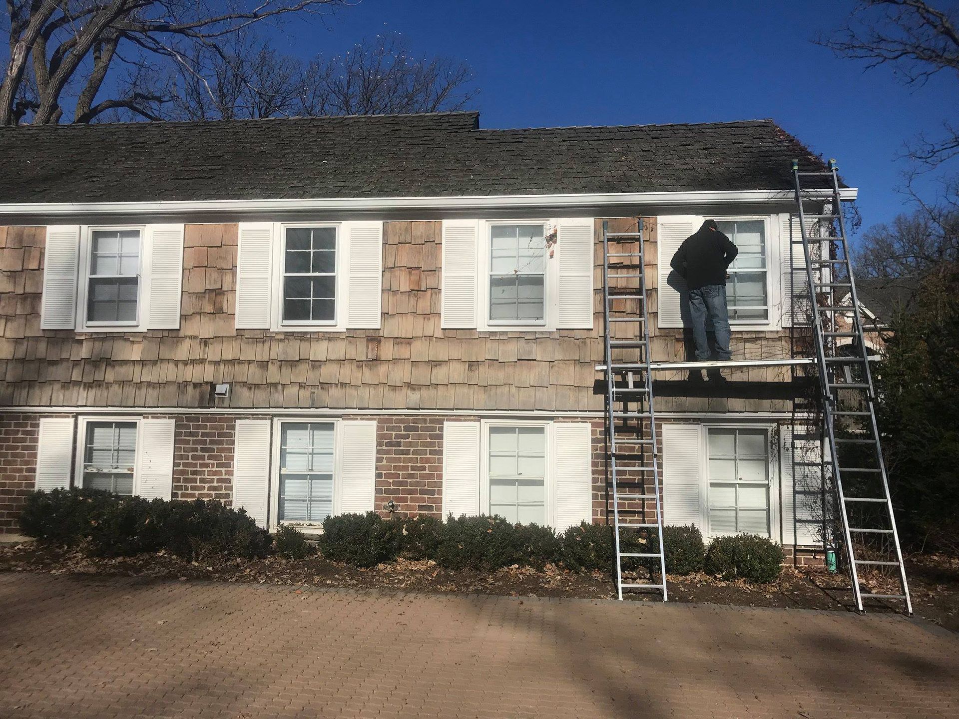 Man on a ladder repairs a house exterior with weathered siding and white shutters.