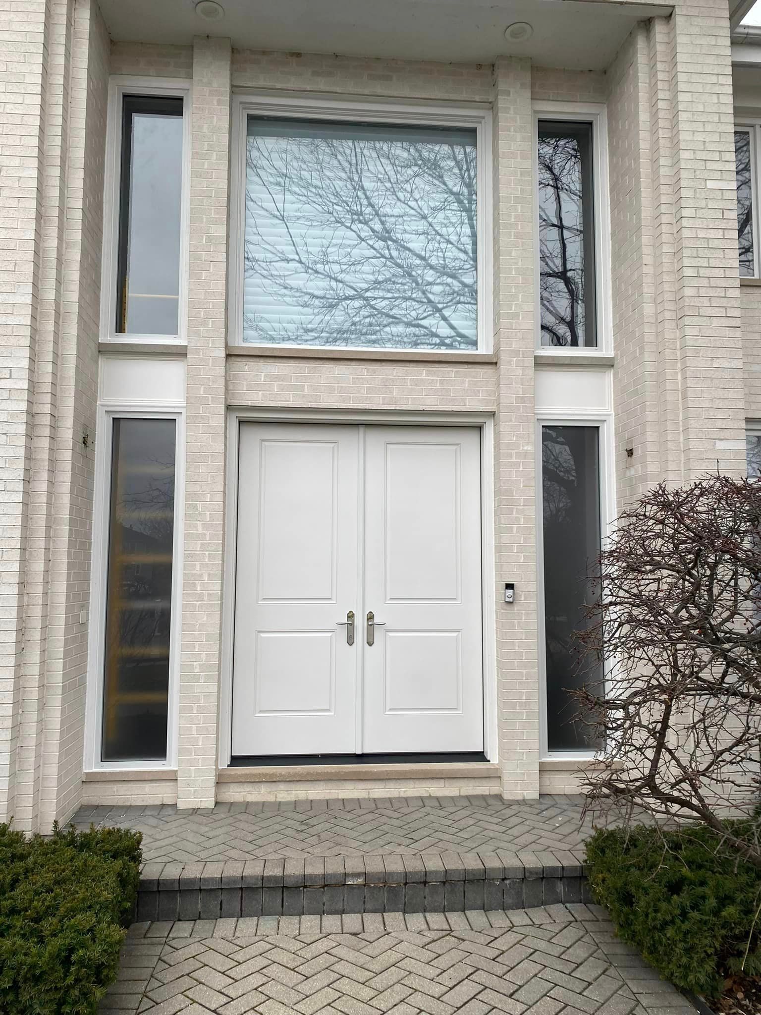 White double doors on a brick house, flanked by tall windows. Paved steps lead to the entrance.