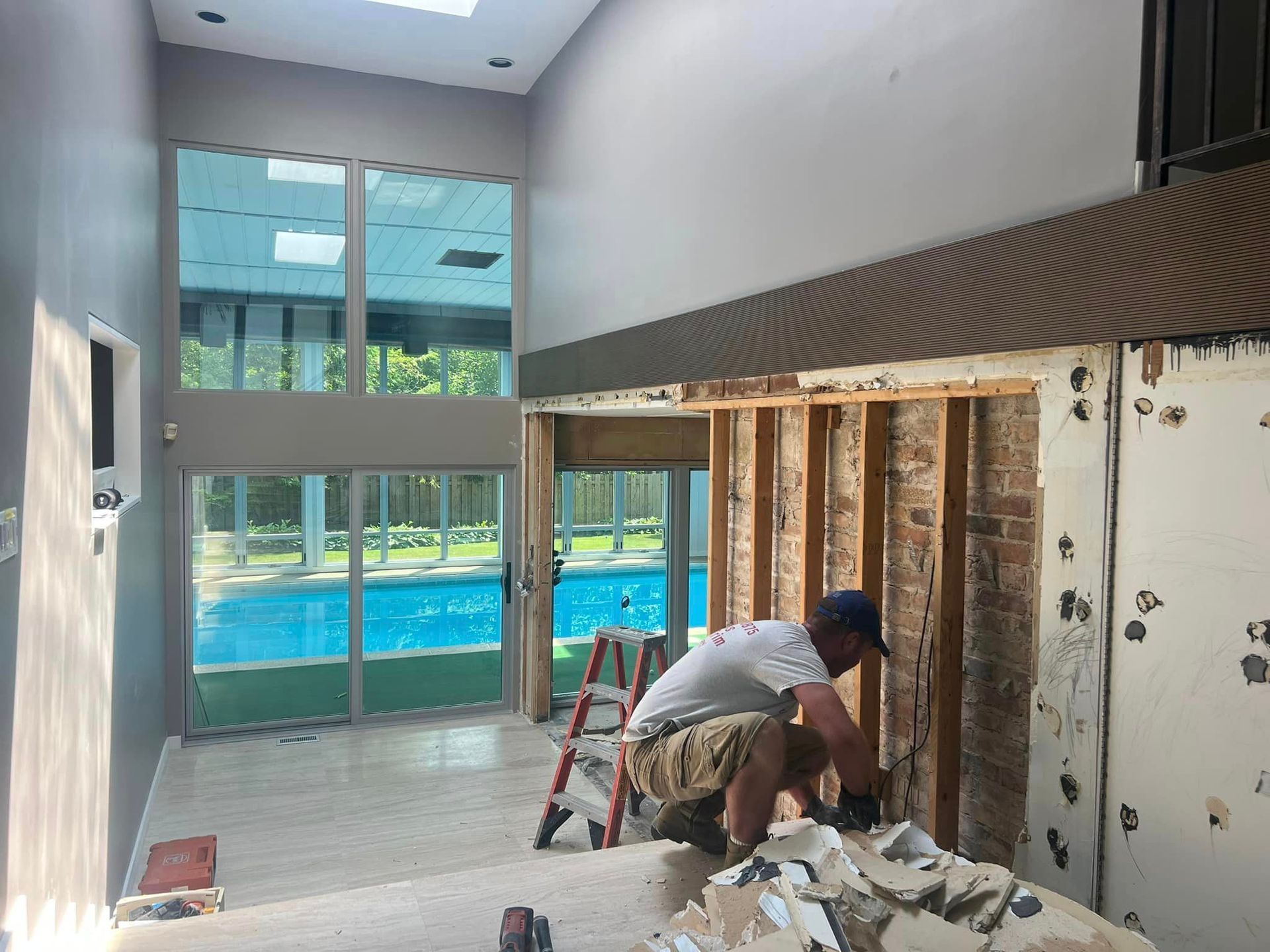 Person demolishing wall in a room with large windows overlooking a pool.
