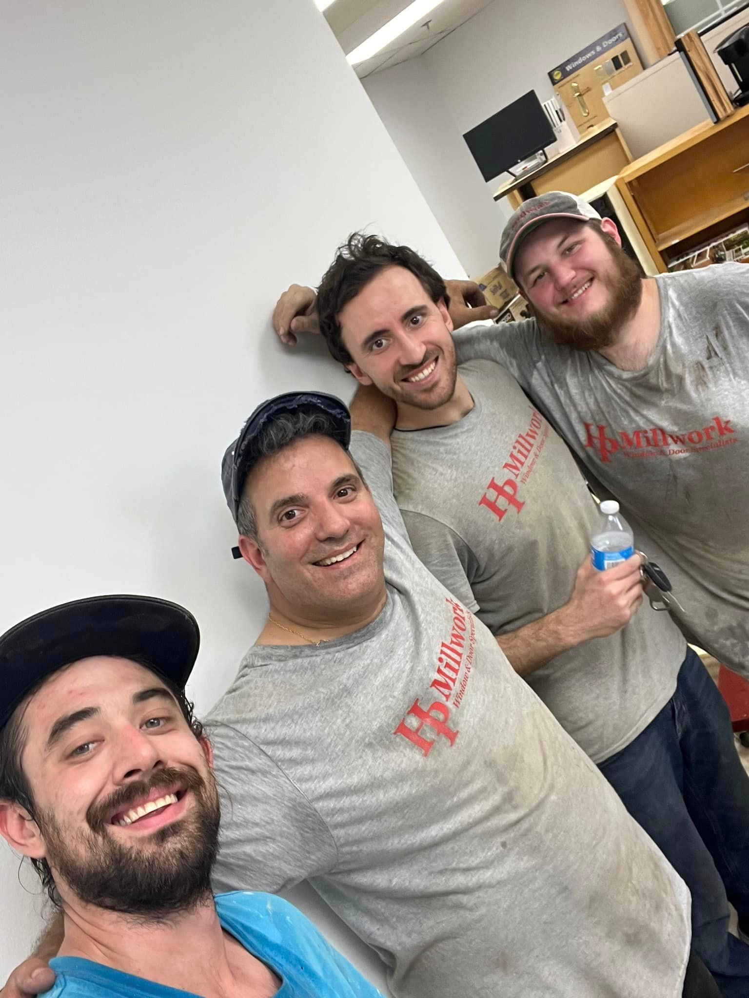 Four men in dusty shirts pose indoors, smiling. One leans on a white wall.
