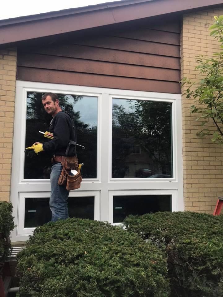Man cleaning windows; house exterior, bushes. Light brick, brown siding, white window frames.
