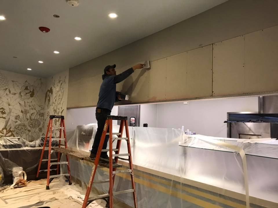 Person on a ladder applies material to wall panels in a room. Construction zone with covered surfaces.