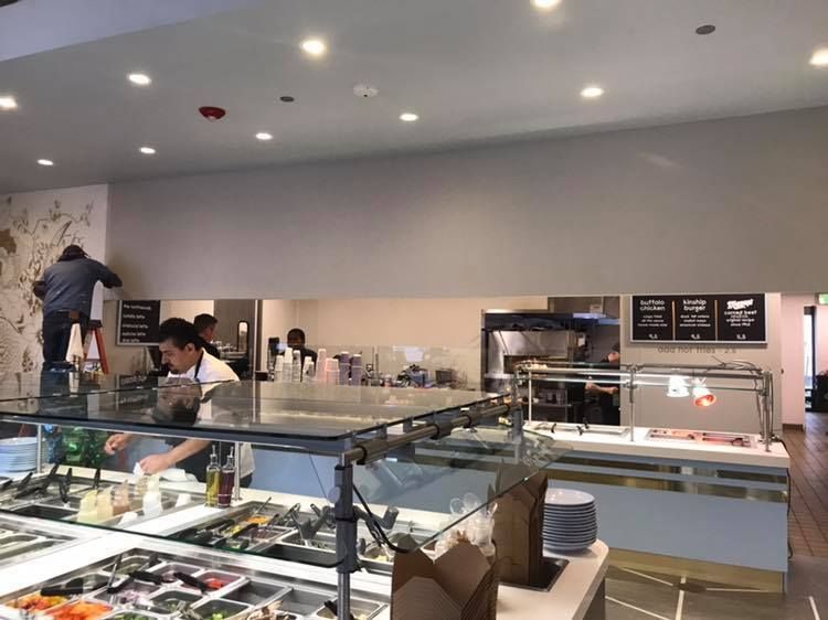 Restaurant interior with workers at salad bar, a person on a ladder working on wall art, and food prep areas visible.