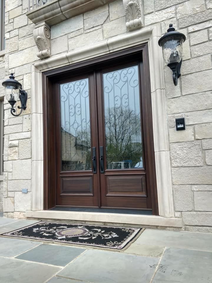 Brown double doors with glass panels, ornate ironwork, and stone facade with two sconces.