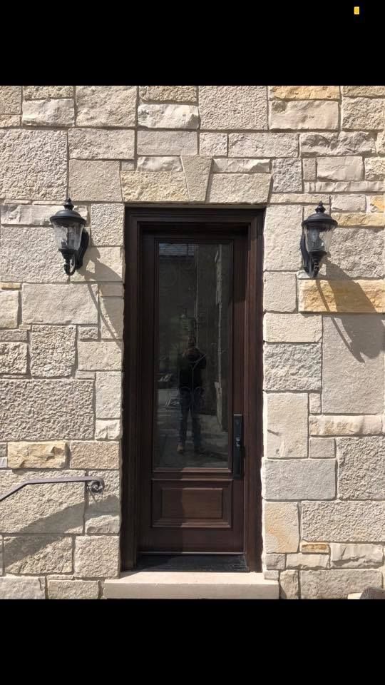 Brown door with glass panel, flanked by black sconces, set in a stone wall.