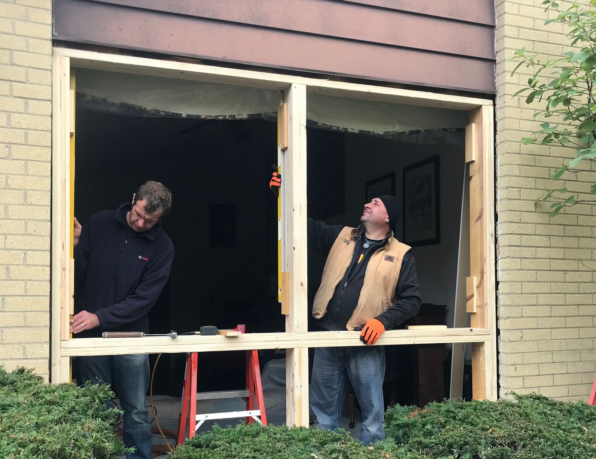 Two people installing a window frame on a brick building; one looks up, other works below.