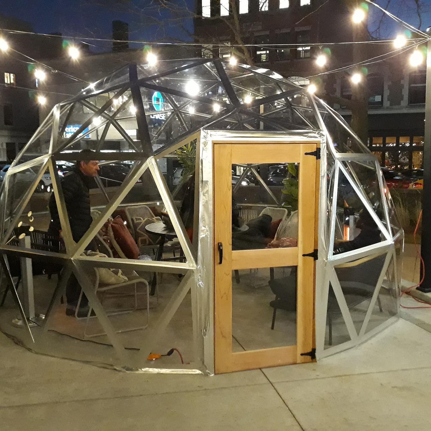 A clear geodesic dome outdoor dining structure. People sit at tables inside, with string lights overhead.