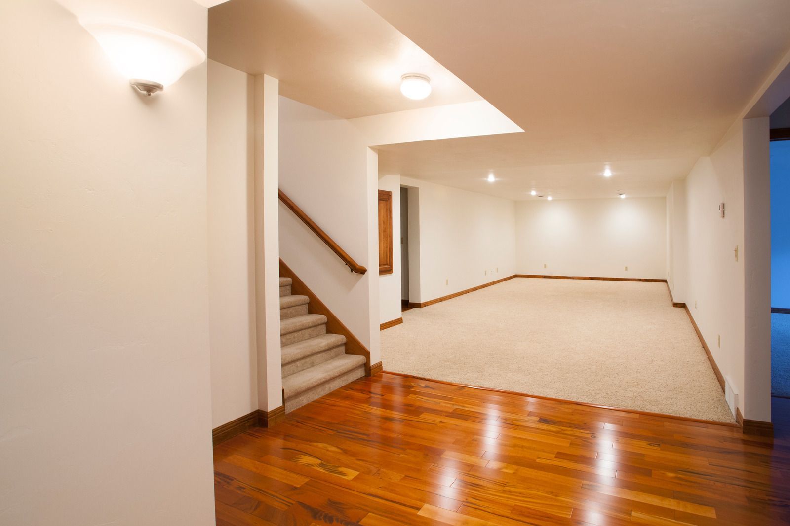 Empty basement with hardwood floors, stairs, and beige carpet. White walls, recessed lighting.