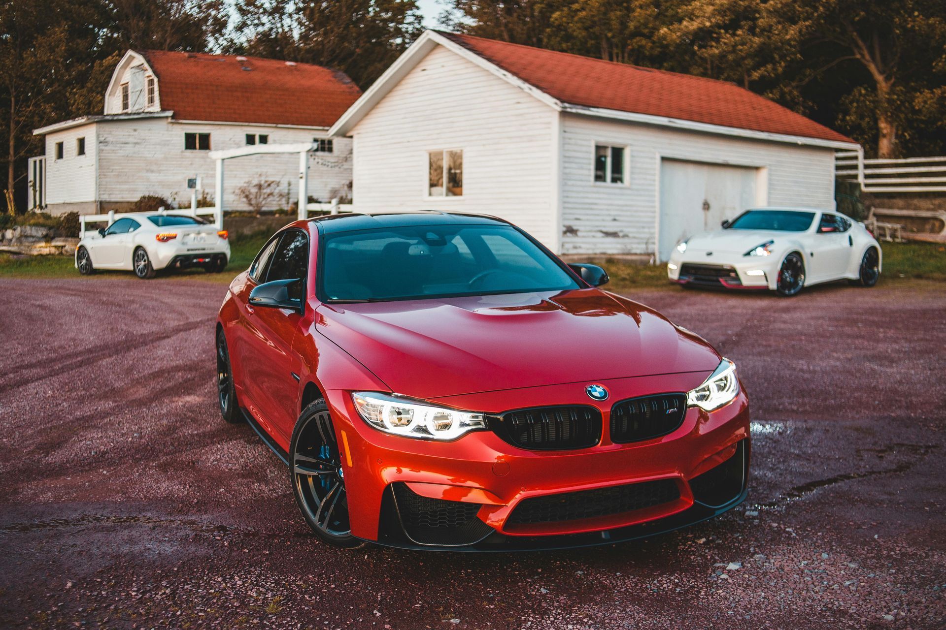 A red BMW M4 parked in a gravel lot with two white sports cars in the background near a white building.