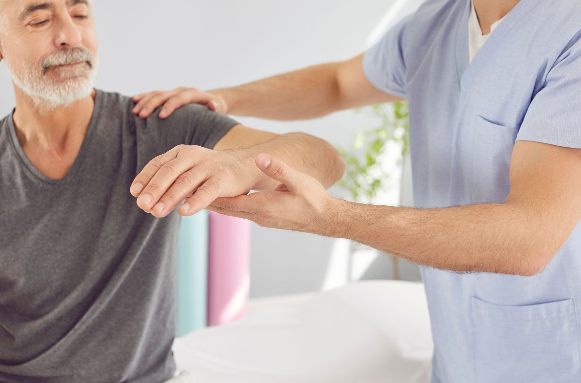 Eine Pflegekraft unterstützt einen sitzenden Patienten sanft bei der Armbewegung in einem hellen Behandlungsraum.