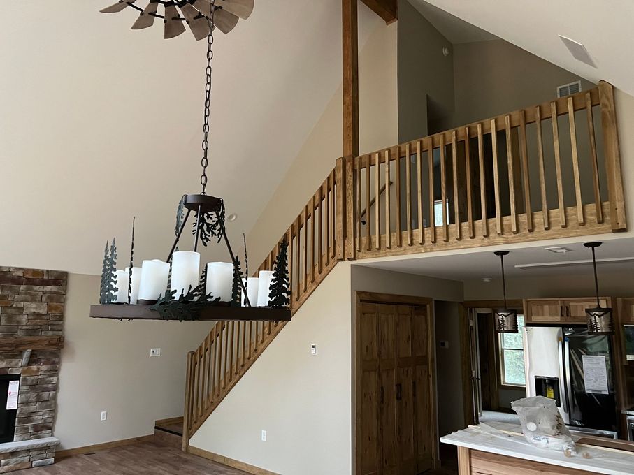 A rustic living space with a high ceiling, a wooden staircase, a metal tree-themed chandelier, and a stone fireplace.