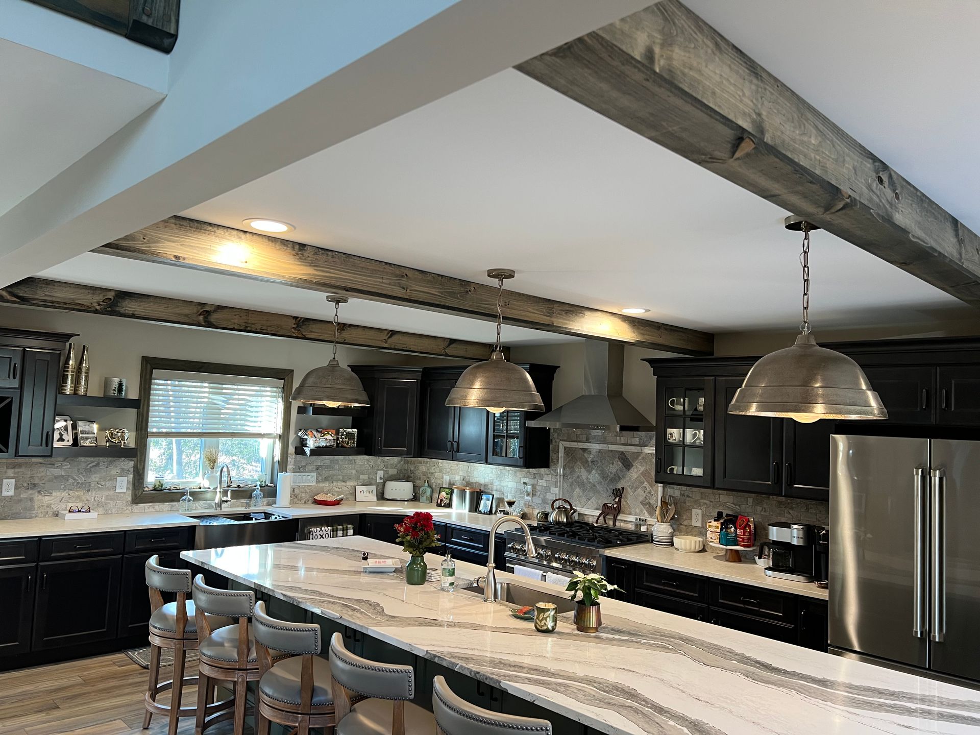 Modern kitchen with a large marble island, dark cabinets, stainless steel appliances, and rustic wooden ceiling beams.